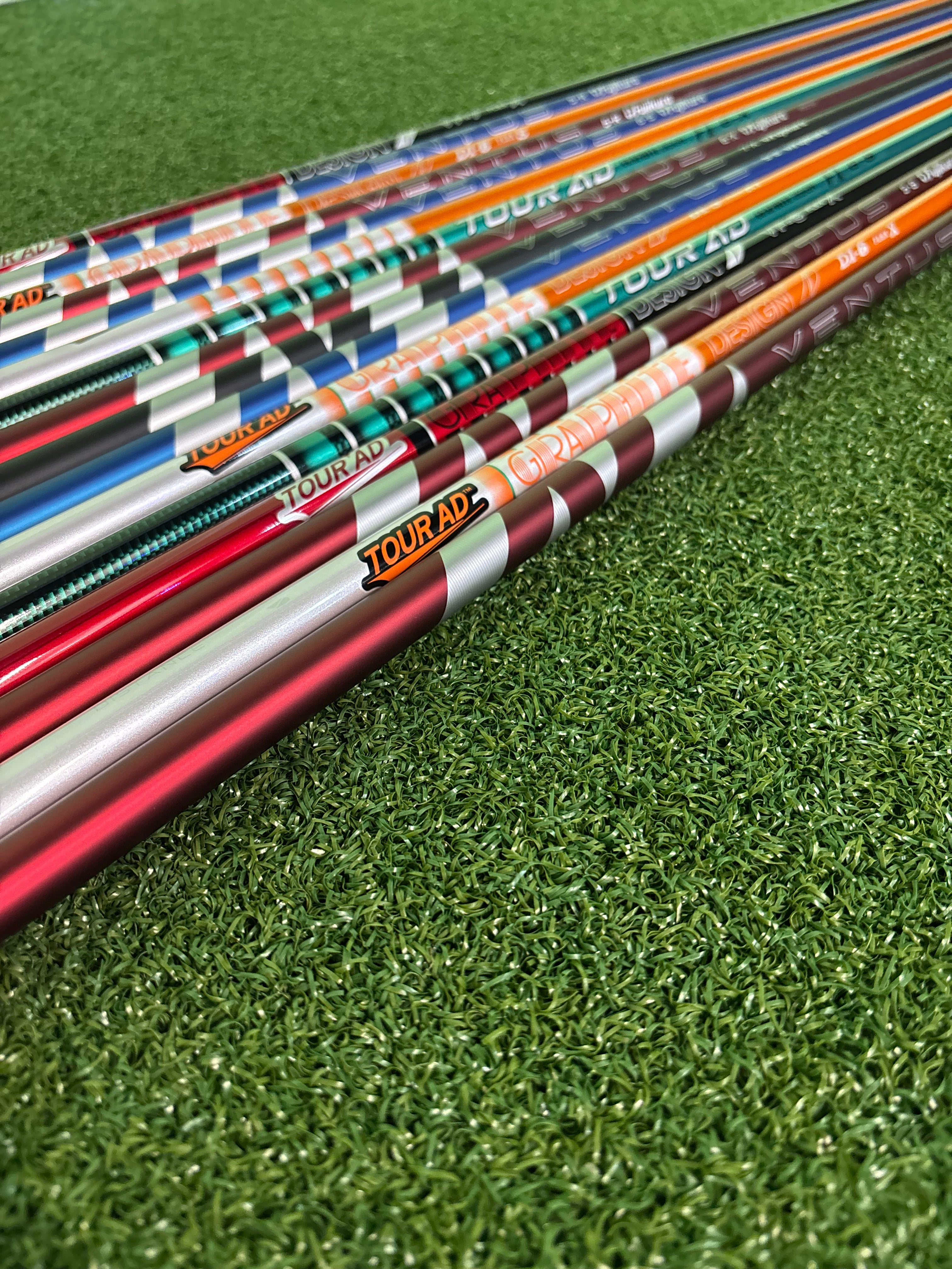 A close-up diagonal shot of several golf shafts resting on green artificial turf. The shafts feature metallic finishes in red, silver, orange, teal, and black, with visible branding including TourAP, TourAD, and TourZA.