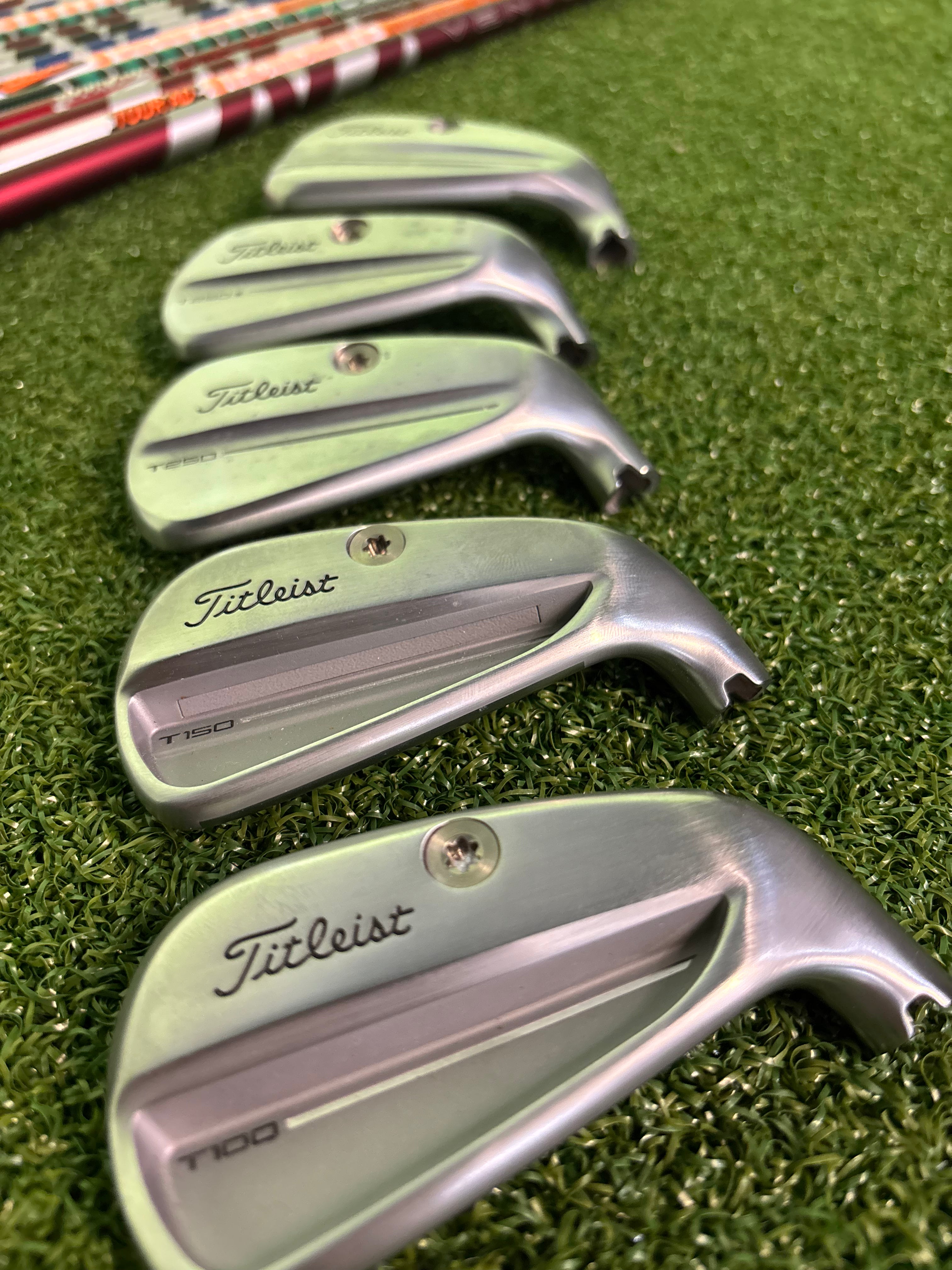 Five Titleist T100 golf iron clubs arranged diagonally on artificial green turf, with the brand name 'Titleist' clearly visible on each club head.
