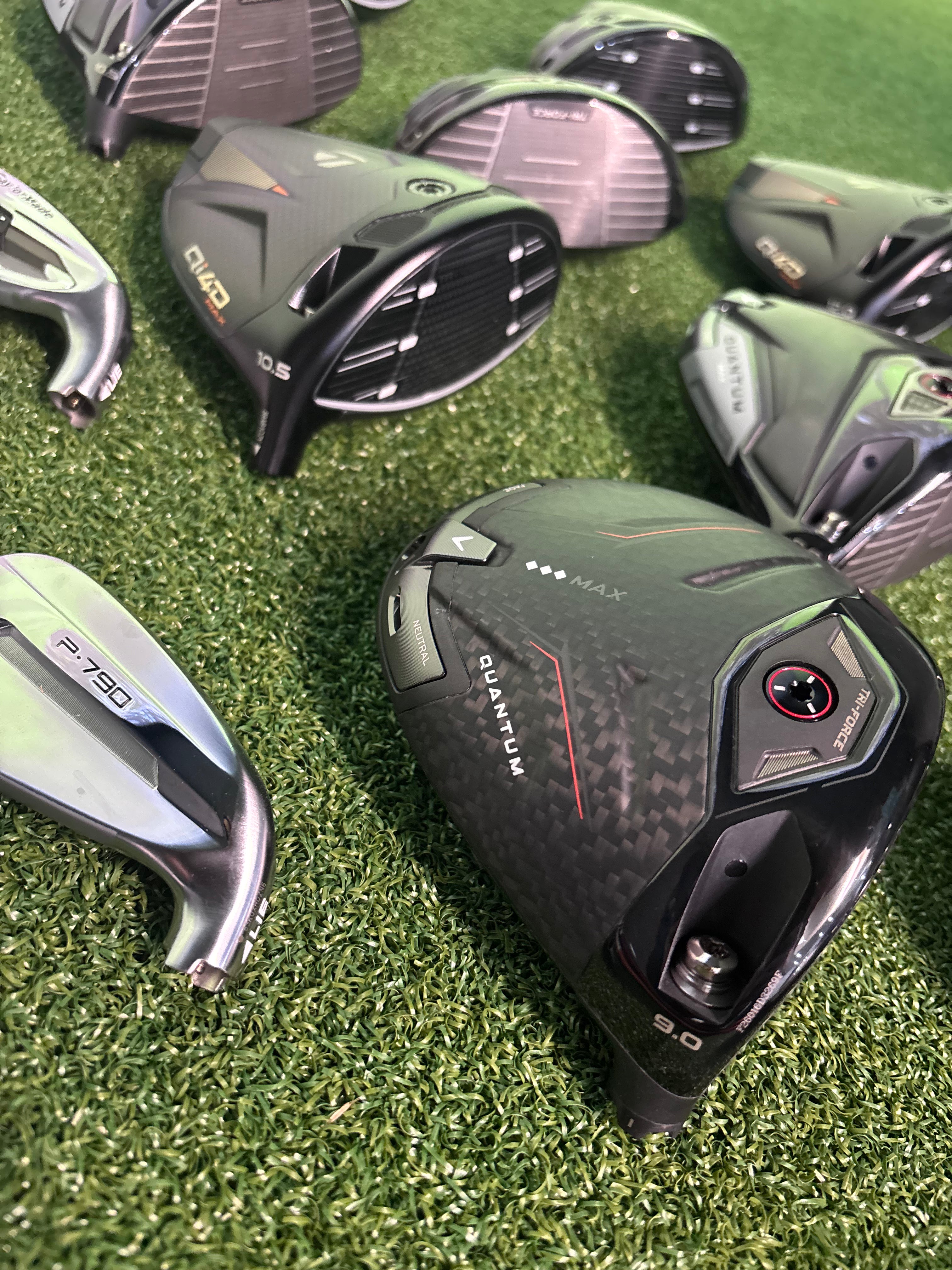 A close-up arrangement of golf clubs on artificial grass, with a black and dark gray carbon-fiber-patterned driver head prominently featured in the foreground. Other visible clubs include a silver P.