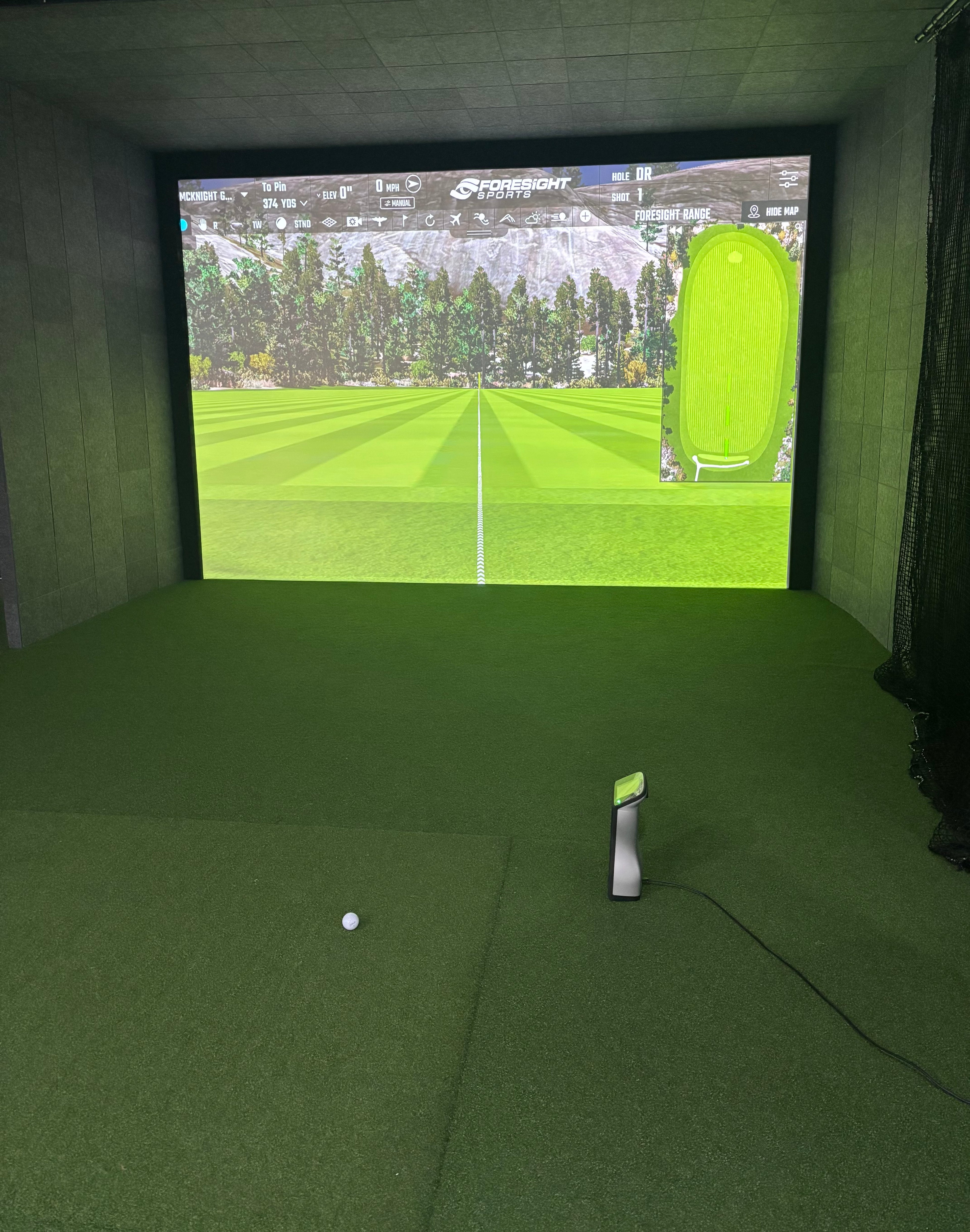 An indoor golf simulator setup with a green turf hitting surface, a golf ball, a launch monitor, and a large projection screen displaying a golf course and green fairway.